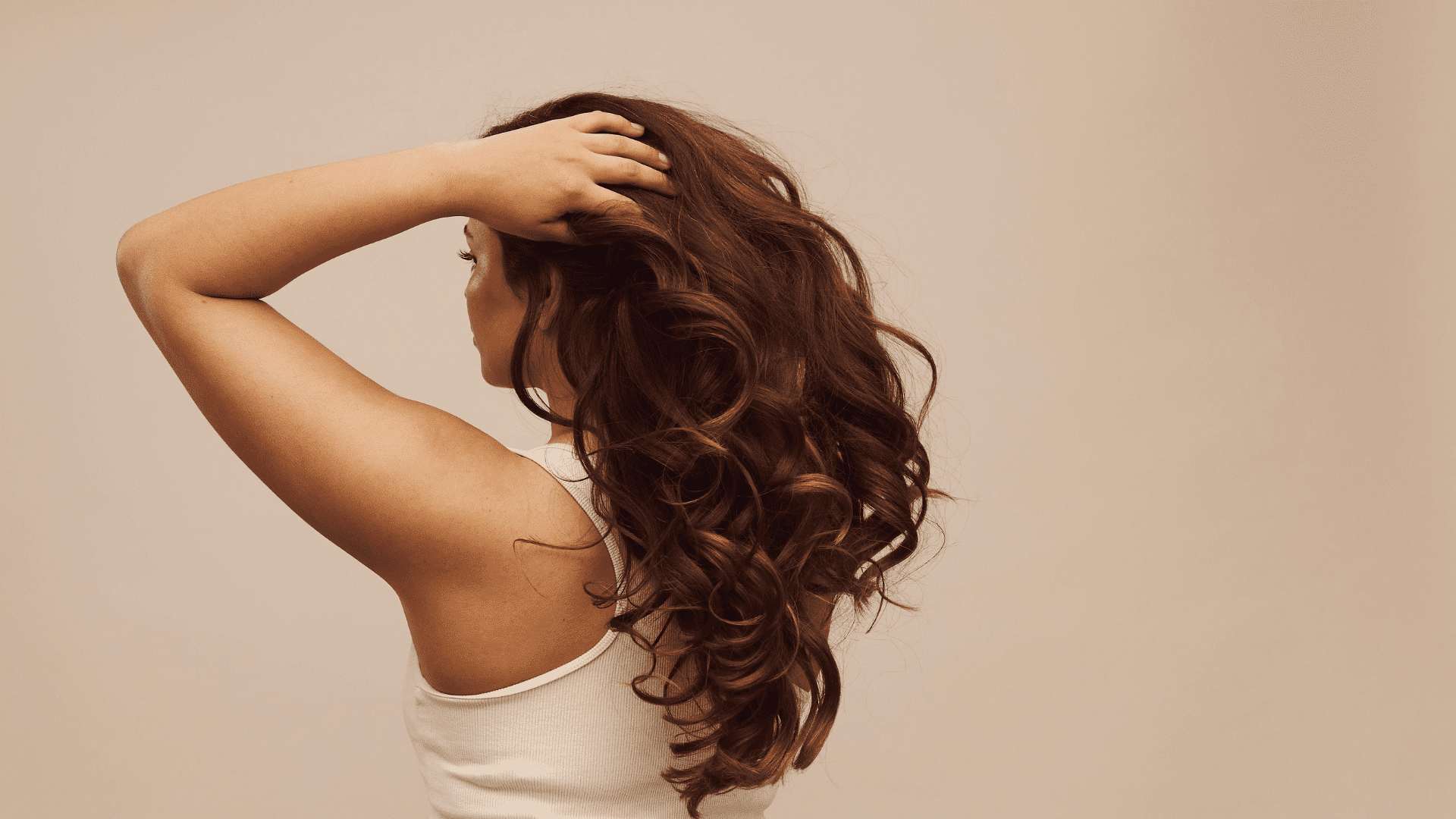Wavy-haired person in a white top, hand on head, facing away against a neutral background.
